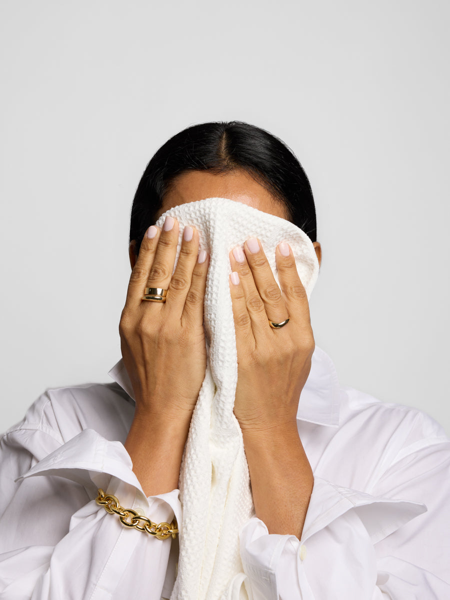 Person wearing a white garment and gold jewelry, covering their face with a white towel against a light gray background.