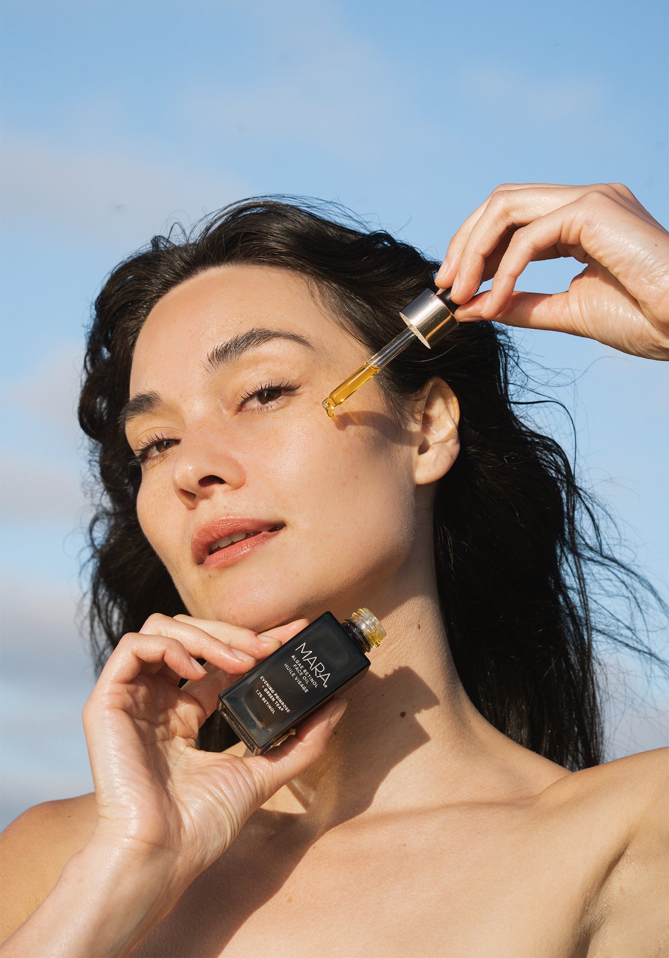 Woman applying Algae Retinol Face Oil with dropper against a clear blue sky