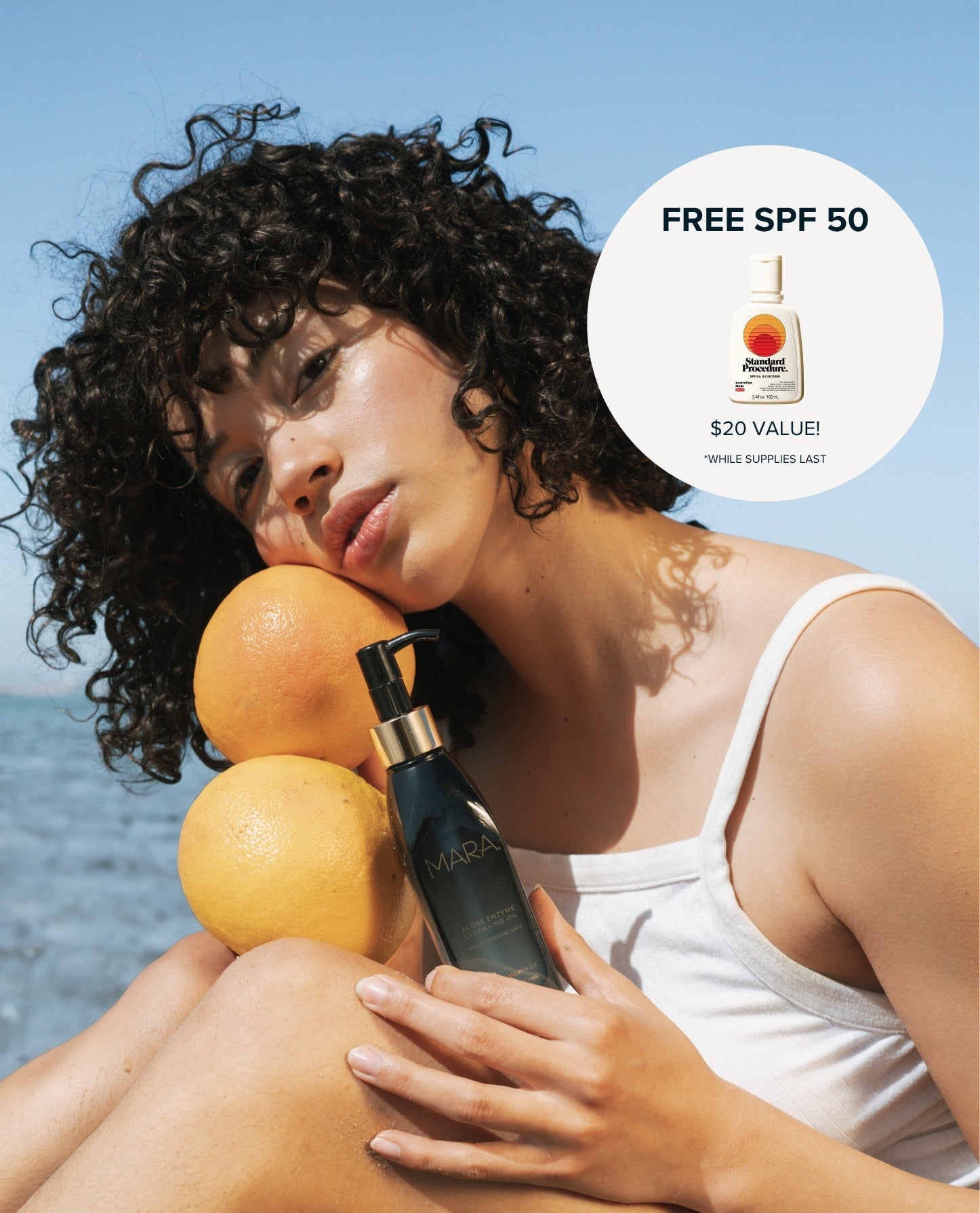 A woman outdoors by the water holds Algae Enzyme Cleansing Oil and two grapefruits