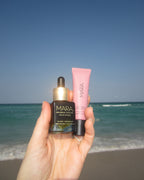 Hand holding MIni Universal Face Oil and Nude Beach Lip Balm at beach