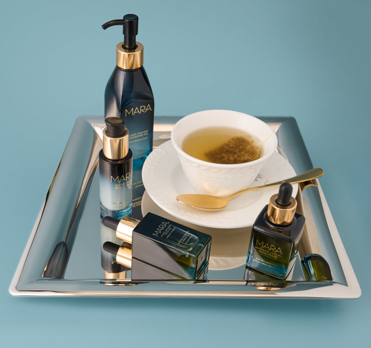 Cleansing Oil, Retinol Oil, Universal Oil, Flower Acid and a cup of tea on a reflective tray with a blue background