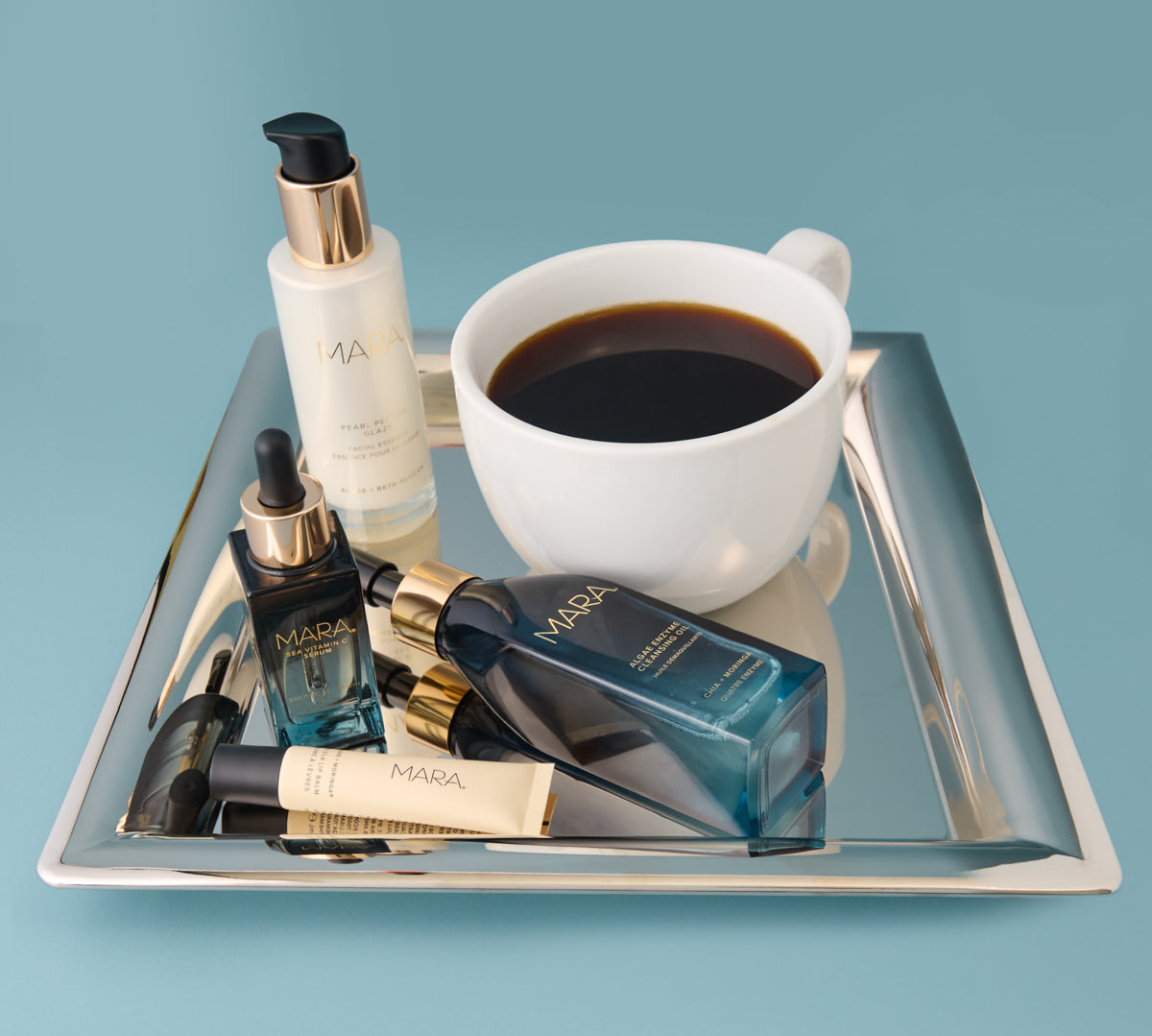 Cleansing Oil, Pearl Peptide, Vitamin C, and Lip Balm and a cup of coffee on silver tray on blue surface