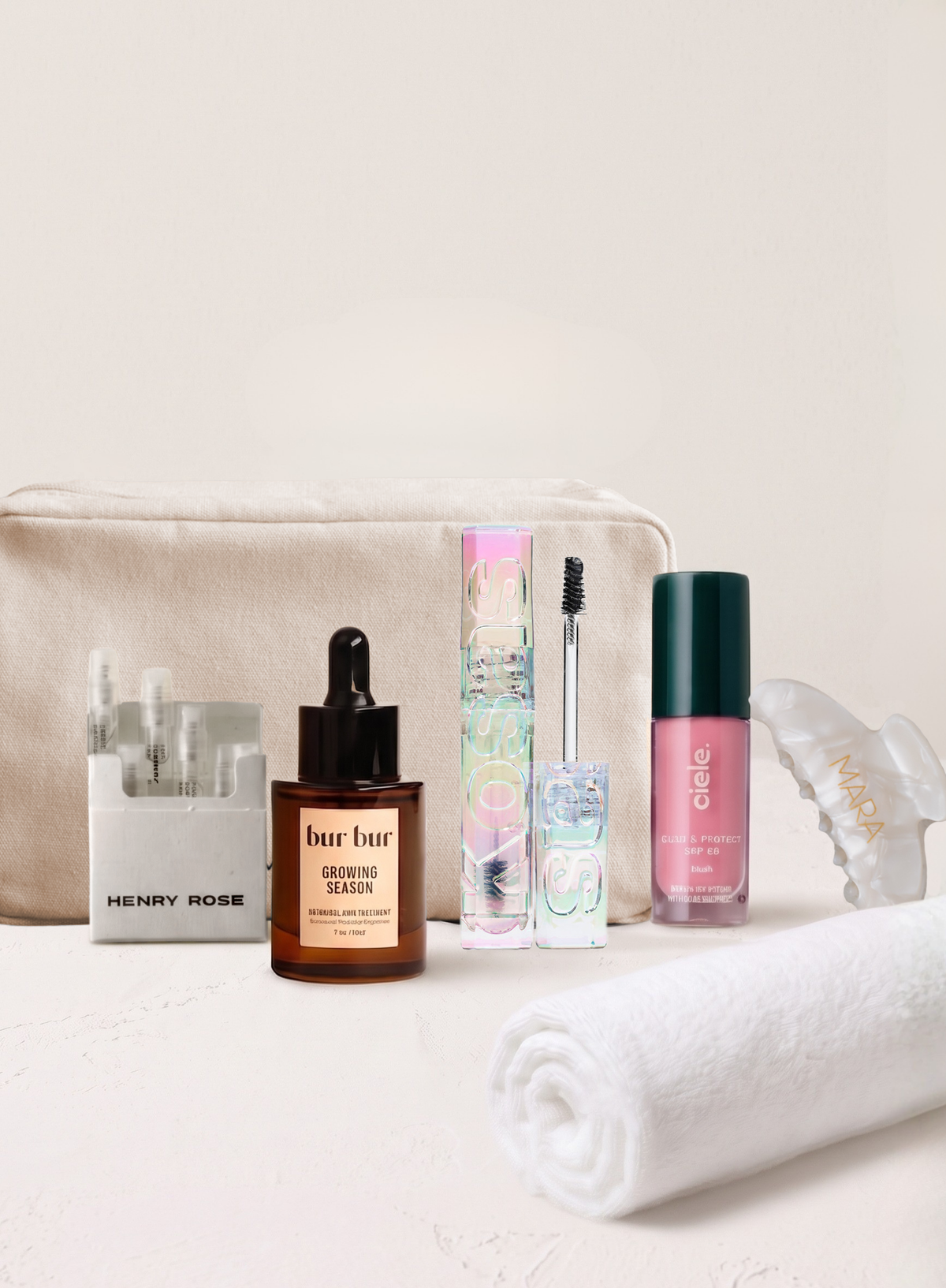 A curated beauty gift set arranged on a light surface against a warm off-white background, including a mara canvas travel bag, bur bur hair oil, kosas brow gel, ciele blush, henry rose discover set, a white rolled towel, and a mara pearl hair claw clip.