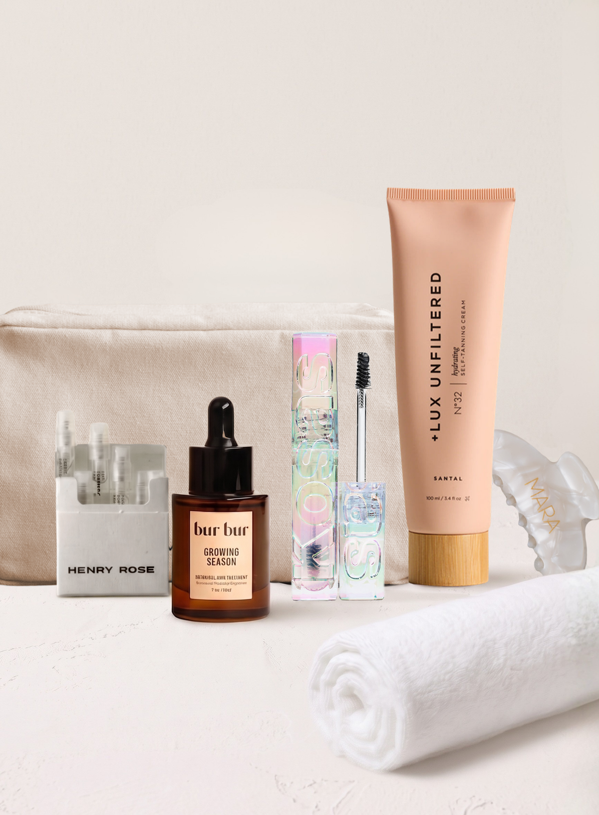 A curated beauty gift set arranged on a light surface against a warm off-white background, including a mara canvas travel bag, bur bur hair oil, kosas brow gel, Lux Unfiltered, henry rose discover set, a white rolled towel, and a mara pearl hair claw clip.