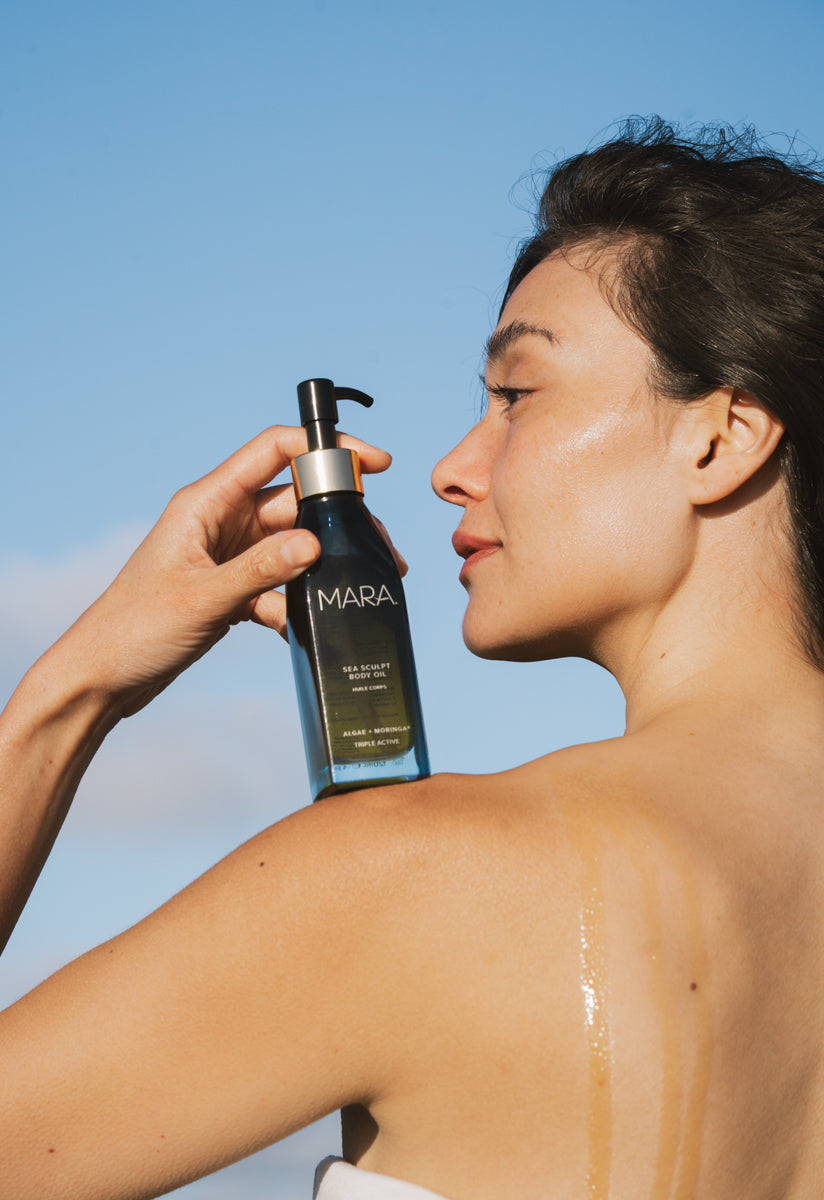 Person holding Sea Sculpt Body Oil to their shoulder against a clear blue sky.