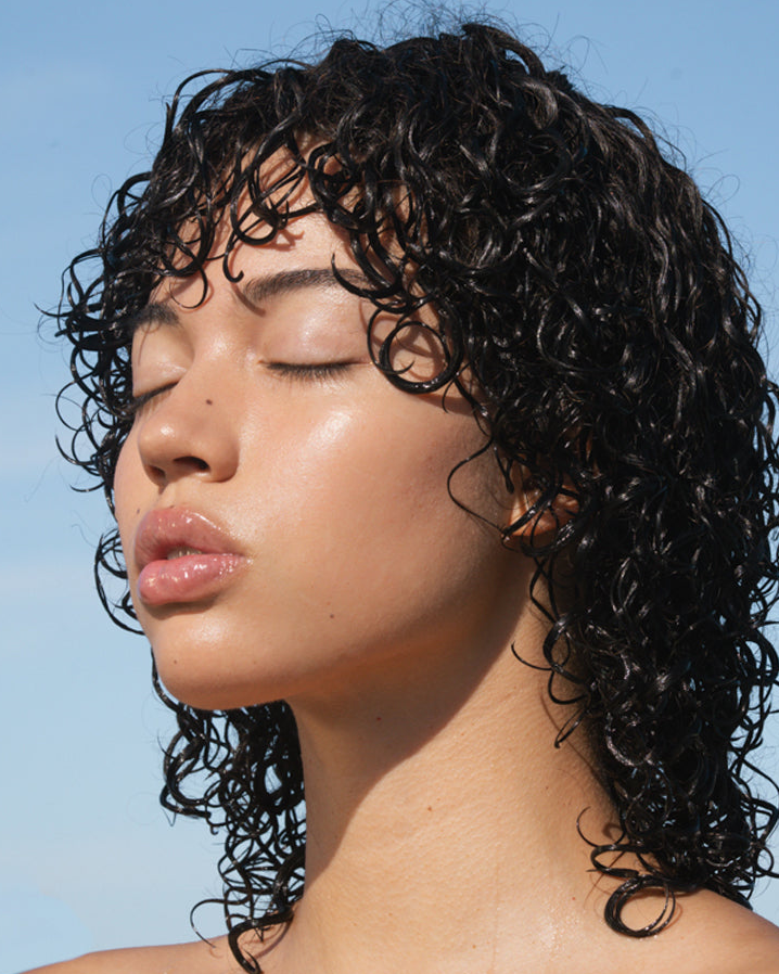 Close-up of a person with closed eyes and wet, curly dark hair against a clear blue sky.