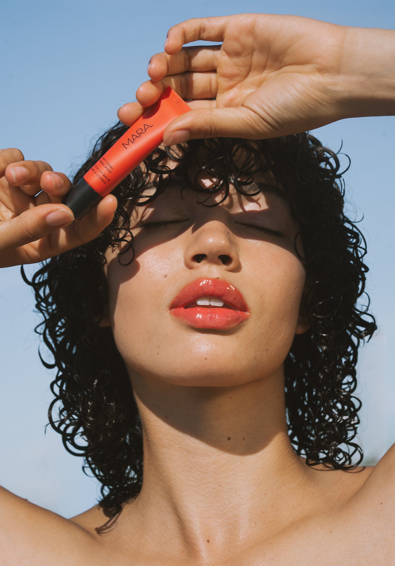 Person wearing and holding Soft Coral Sea Silk Lip Balm above her head against a clear blue sky