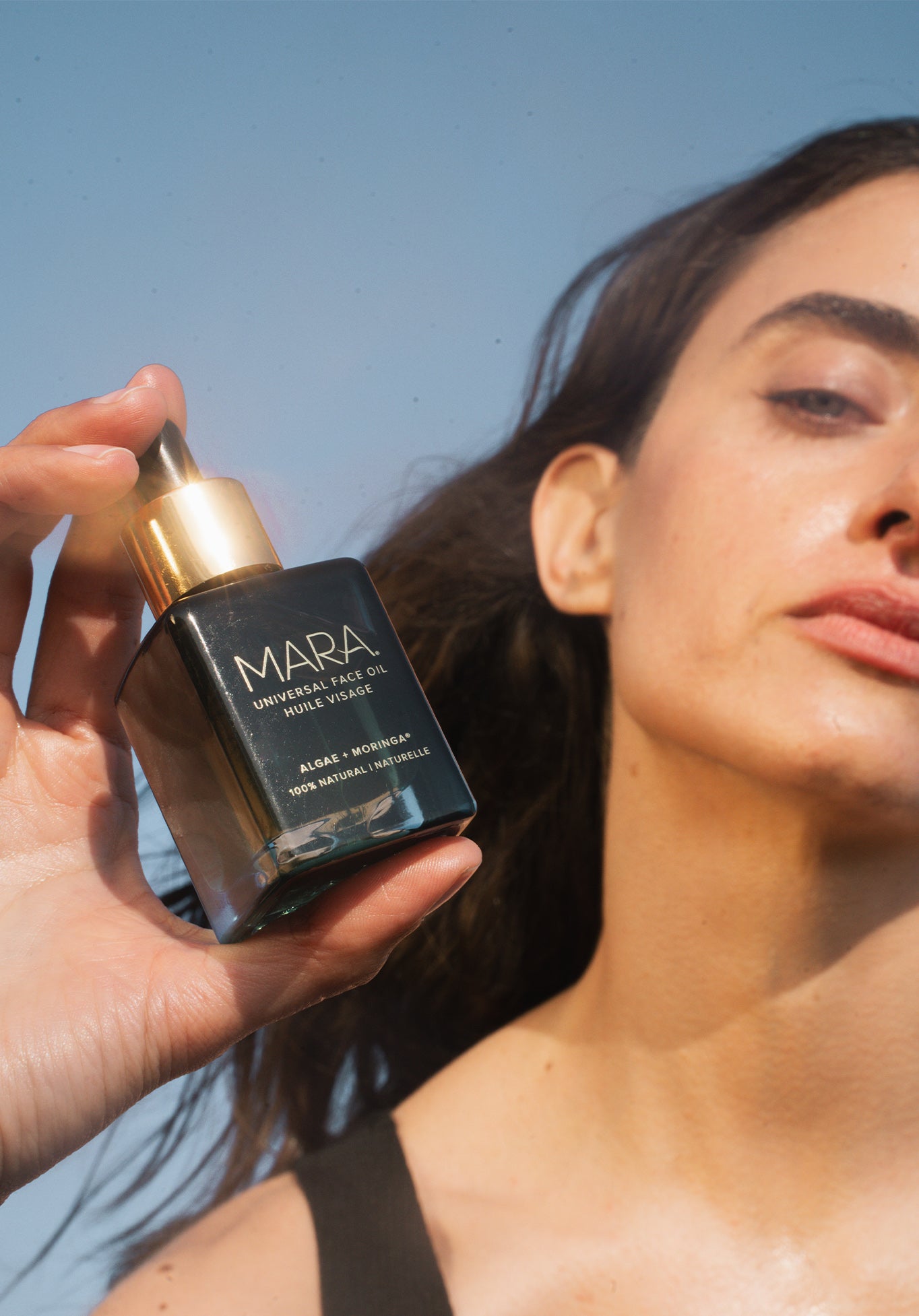 Person holding a bottle of Universal Face Oil in front of her against a clear blue sky