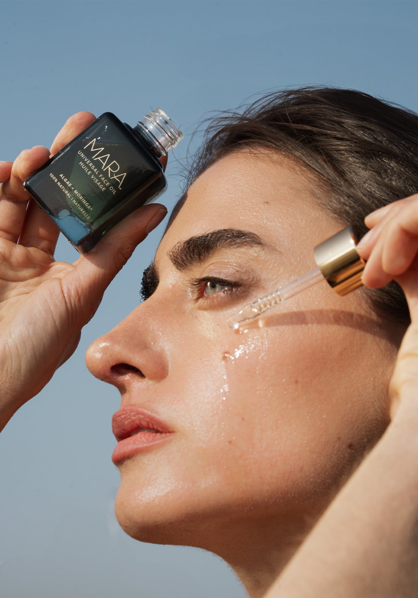Person applying Universal Face Oil with a dropper against a clear blue sky