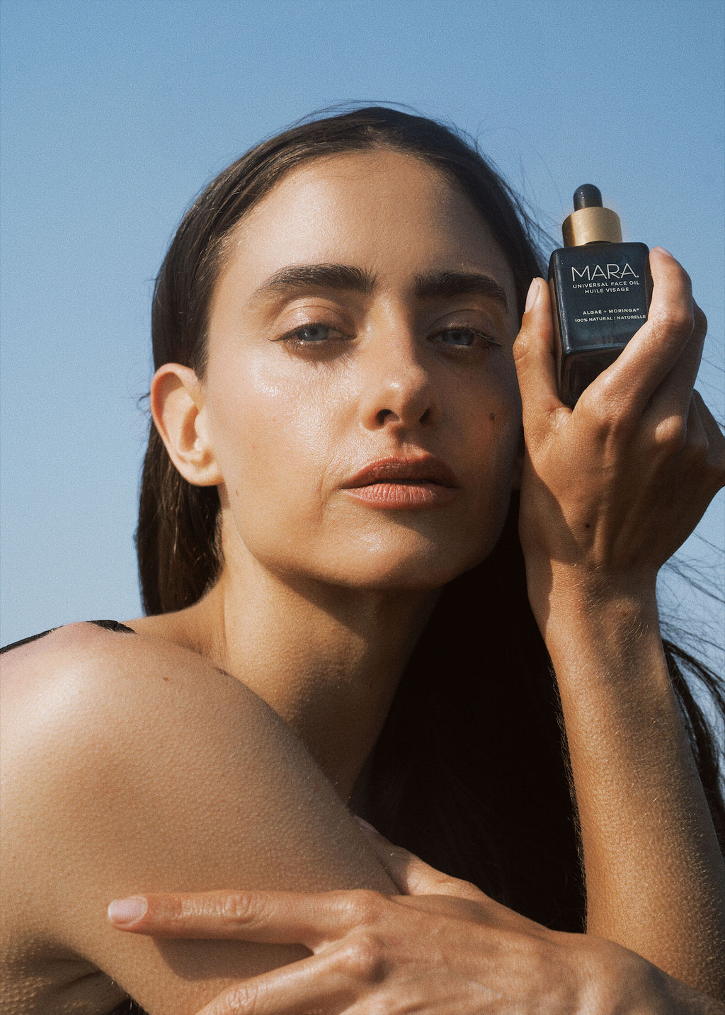 Woman holding a bottle of Universal Face Oil against a clear blue sky.