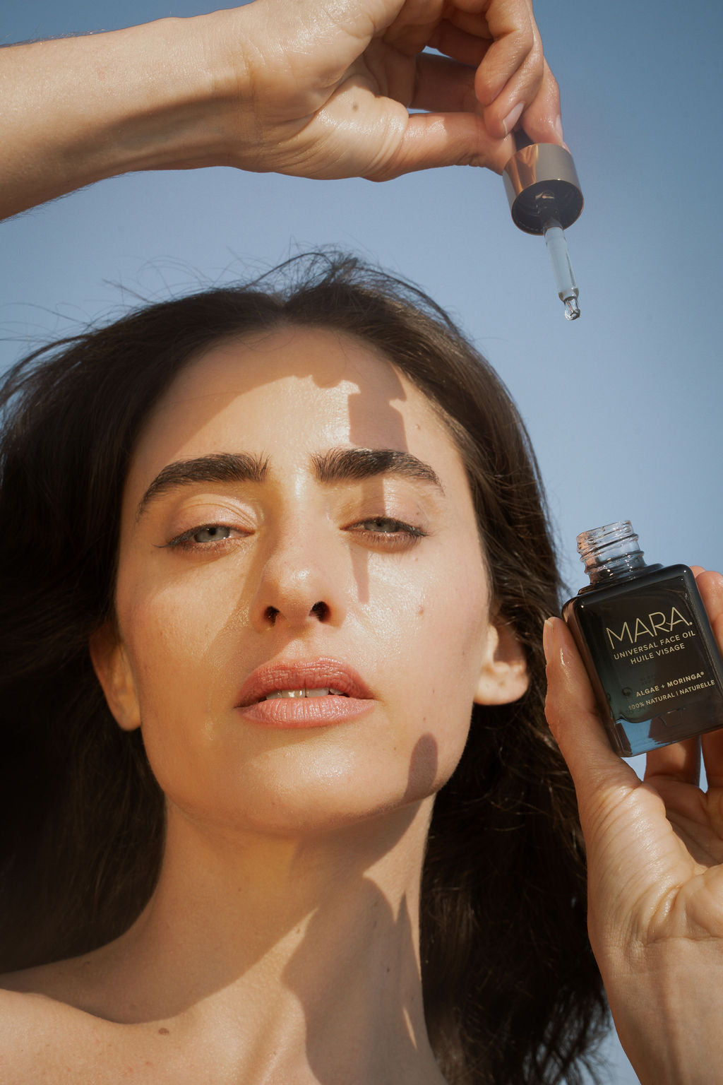 Person applying Universal Face Oil with dropper against a clear blue sky.